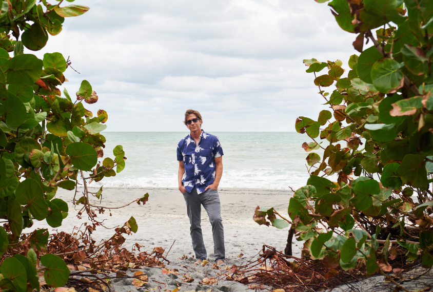 Gavin Rumbaugh stands on a beach alone, framed on the left and right by large, leafy, green plants.