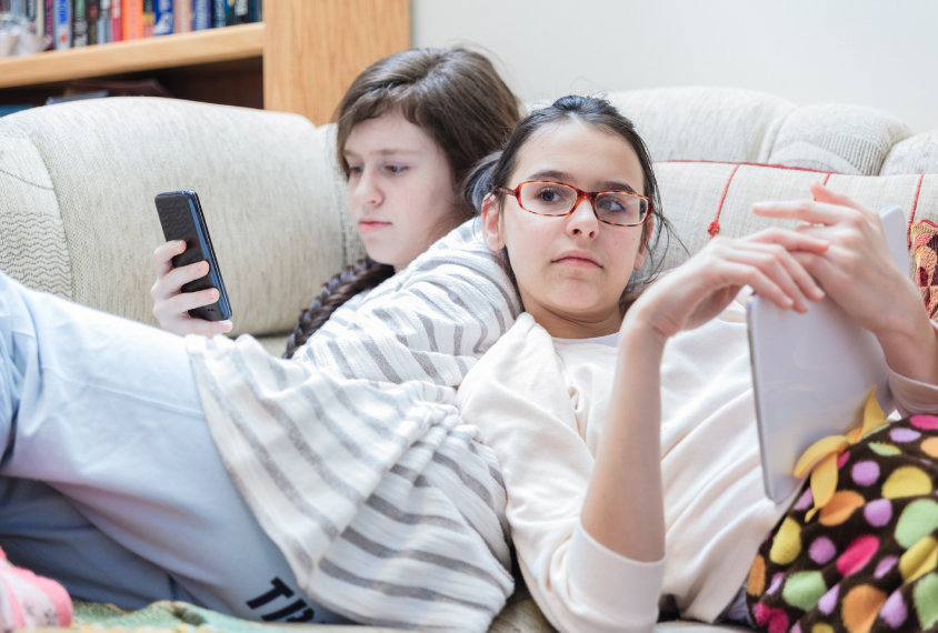 Two teenage girls sit next to each other on a couch, one looking at a cell phone while the other uses a tablet computer.