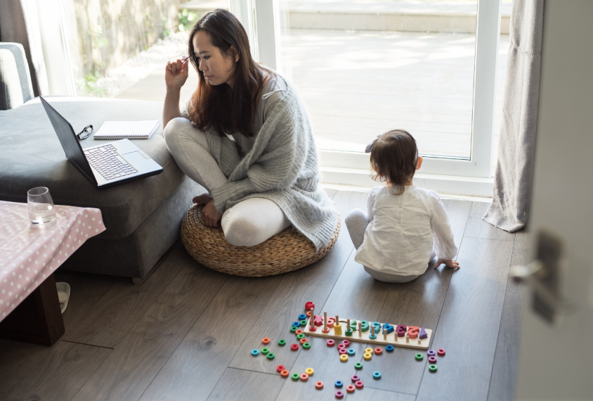 A woman sits and works at a laptop while her daughter sits next to her and looks out of a glass door.