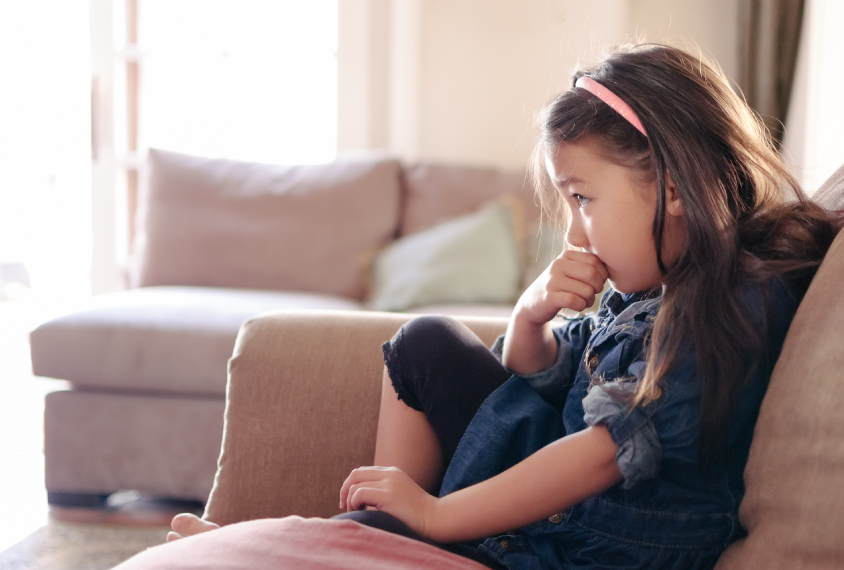A young girl sits on the couch biting her thumb.