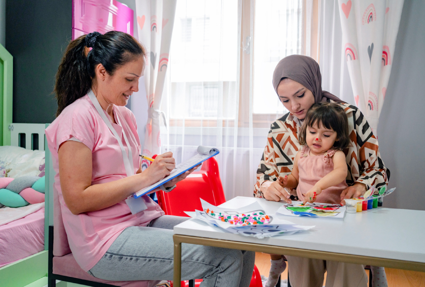 Photograph of a doctor taking notes on a toddler who is playing with their mother.