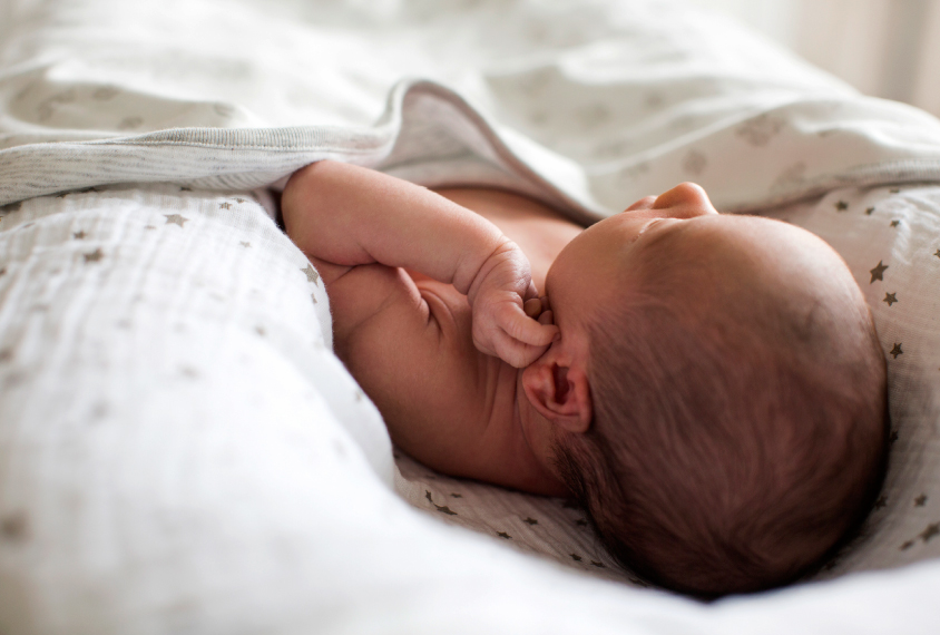 Closeup photograph of an infant lying in a bassinet under white sheets with their left hand lifted close to their ear.