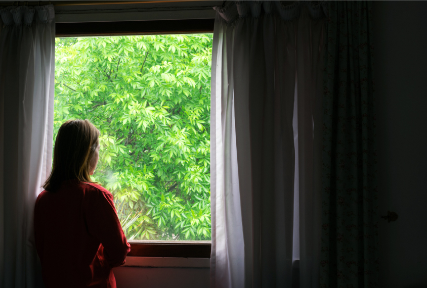 Photograph of a woman in a dim room looking out her window.