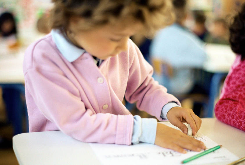 Out-of-focus photograph of a young girl sitting at a desk using an eraser.