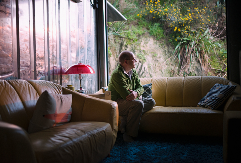 Photograph of a white man in a green sweater sitting on a yellow sofa near a large window.