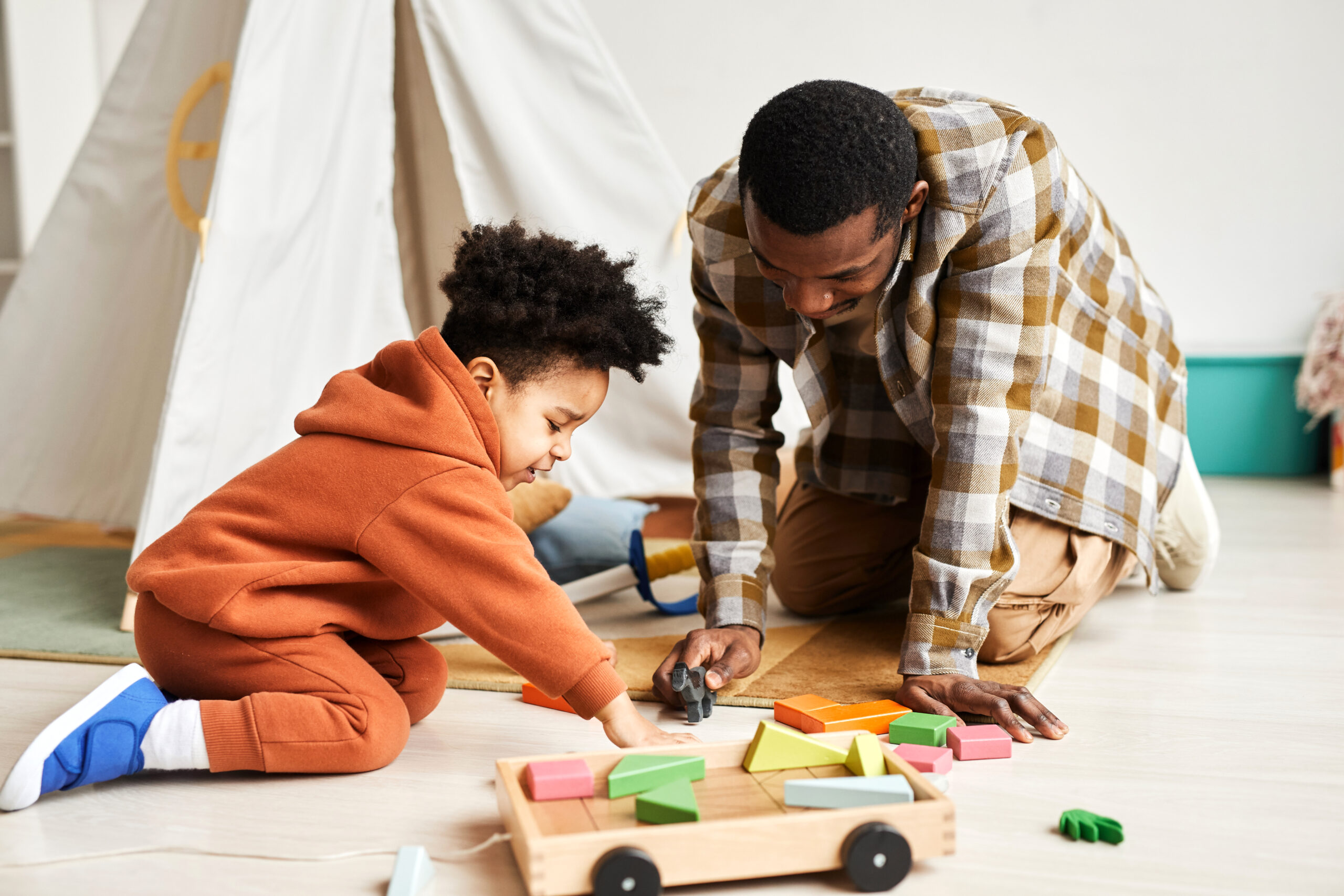 A father and son play with a wooden toy horse on the floor.