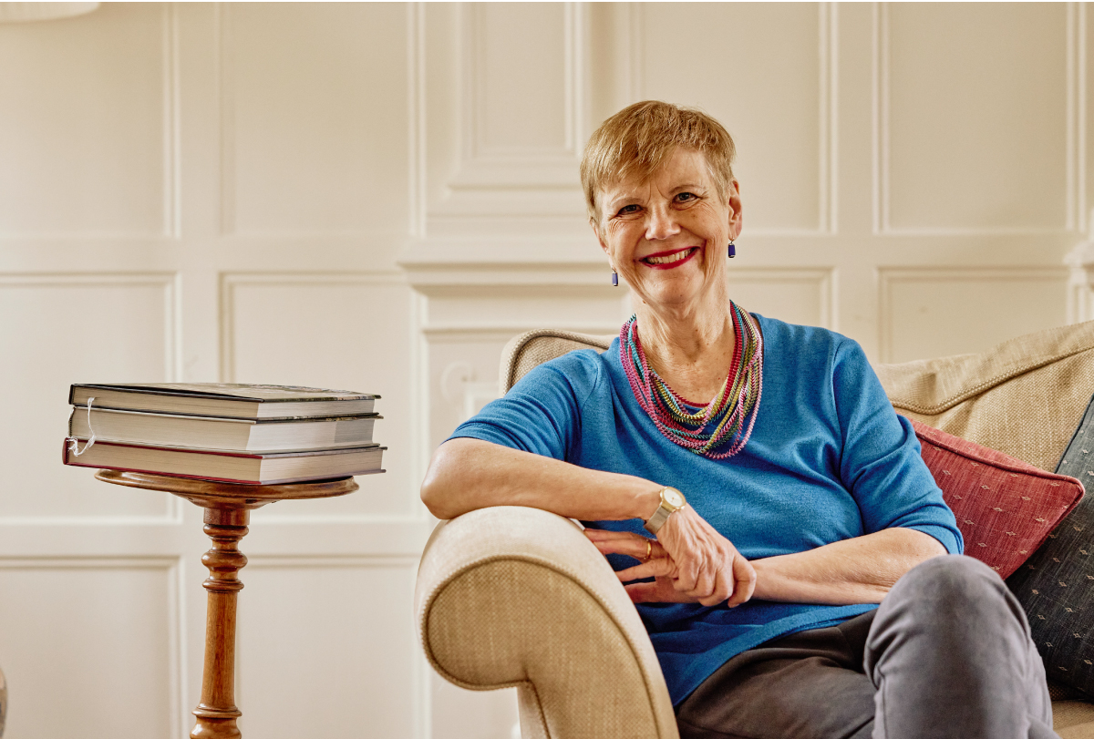 Dorothy Bishop sits on a couch next to a stack of books on a small, circular table.