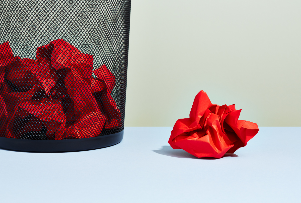 A wadded up piece of red paper sits next to a wastebasket filled with similar pieces of crumped red paper.