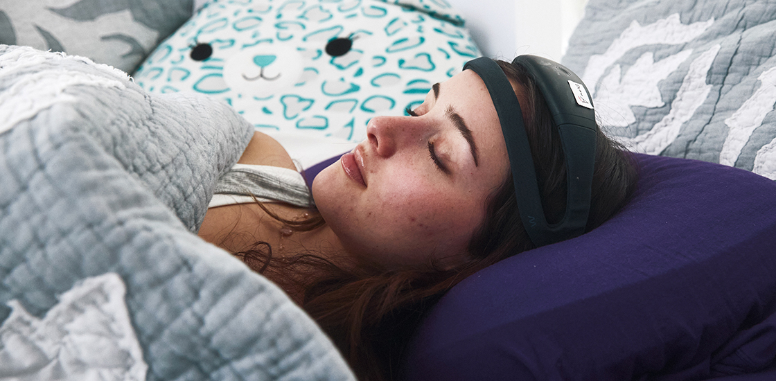 A young woman sleeps at home wearing a Dreem headband and surrounded by stuffed animals.