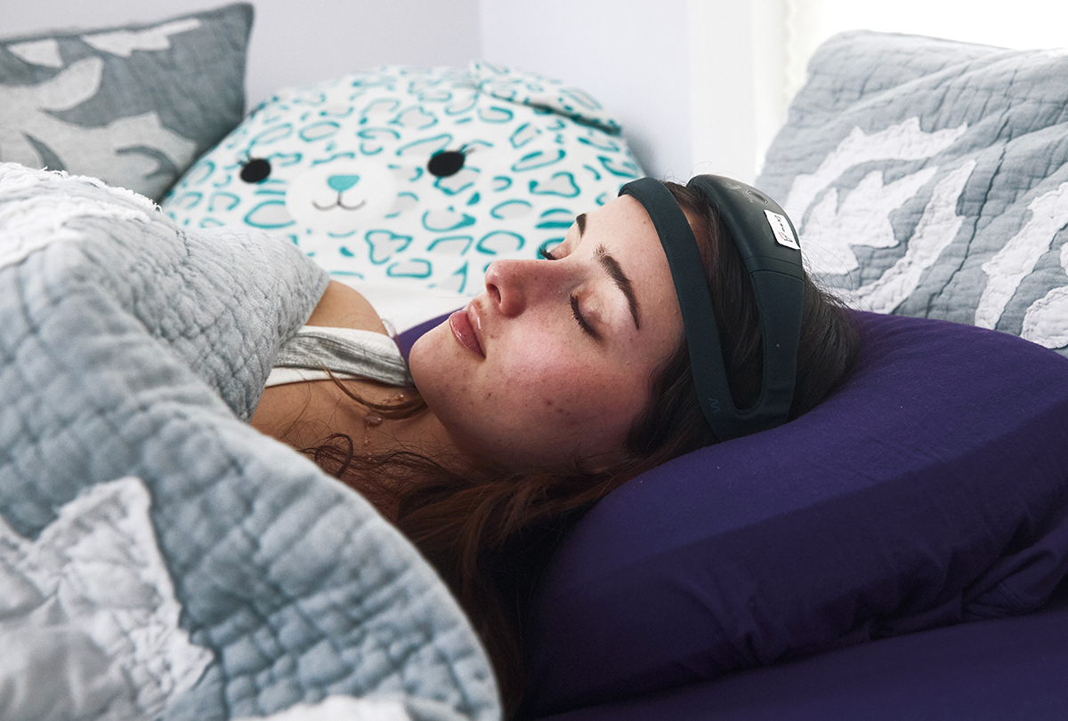 A young woman sleeps at home wearing a Dreem headband and surrounded by stuffed animals.