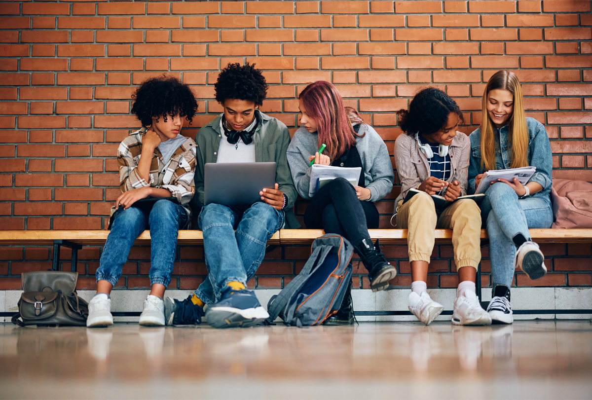 Five high-school-aged students sit on a bench looking at notebooks and a laptop.