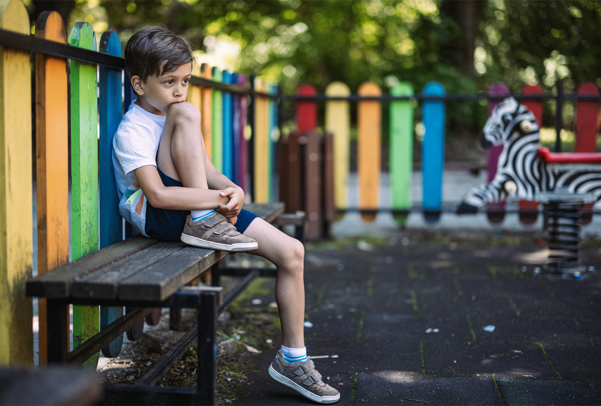 A young boy sits alone on a bench in a playground.