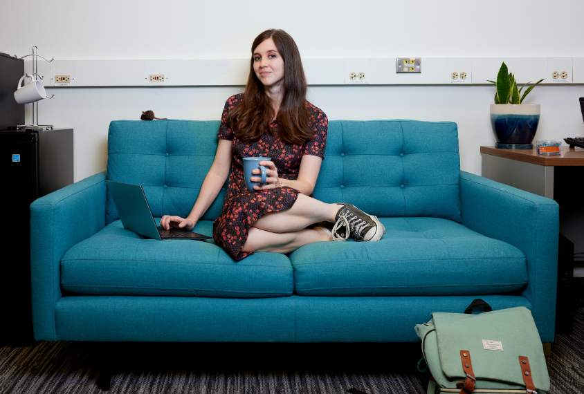 Ann Kennedy sits on a turquoise couch. She holds a light blue mug in her left hand and her right hand is placed on her laptop's keyboard.