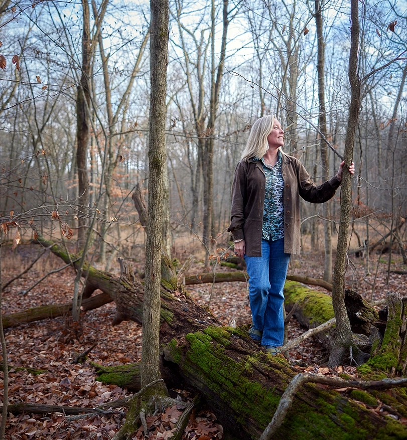 Susan Masino standing on a mossy log among the trees.