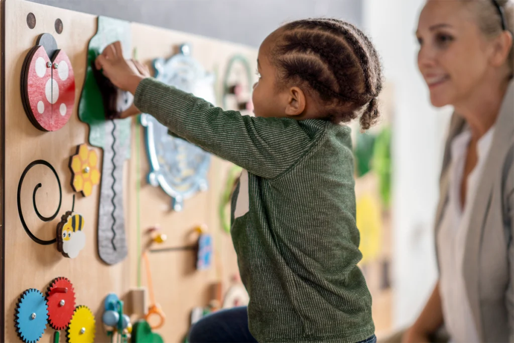 A preschool-age child plays with a vertical sensory board to which colorful moving objects are attached.