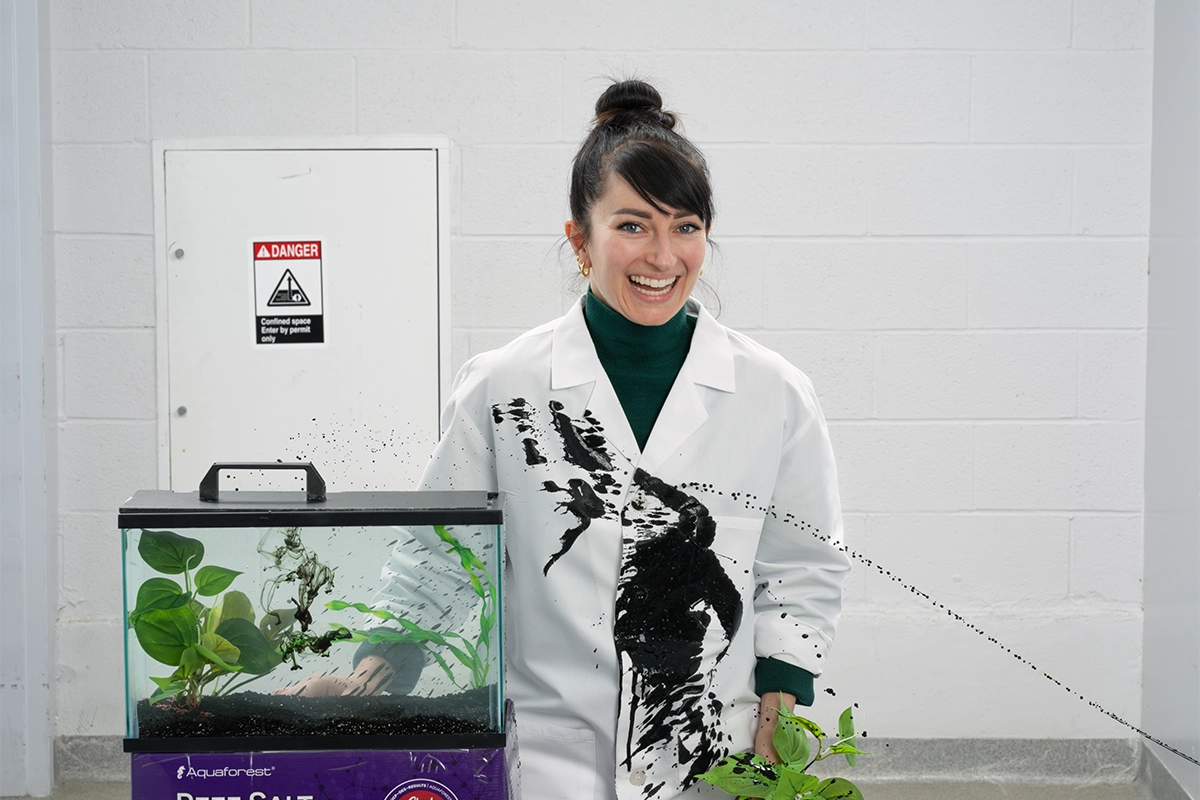 Portrait of scientist Tessa Montague standing next to an aquatic tank. A spray of black ink shoots onto her lab coat from off-camera.