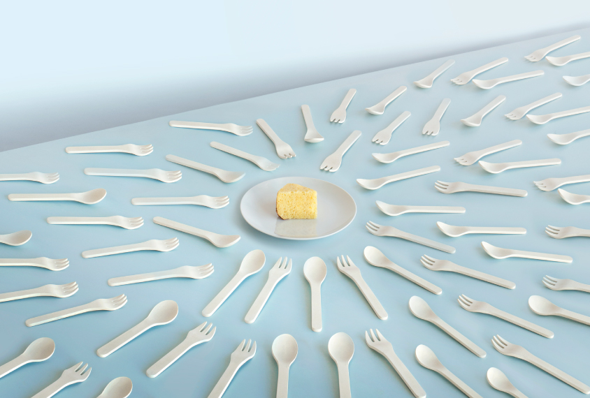 A plate with a slice of yellow cake sits on a blue plate on a light blue table cloth, surrounded by spoons and forks. This image places an emphasis on food and ritualized behaviors.