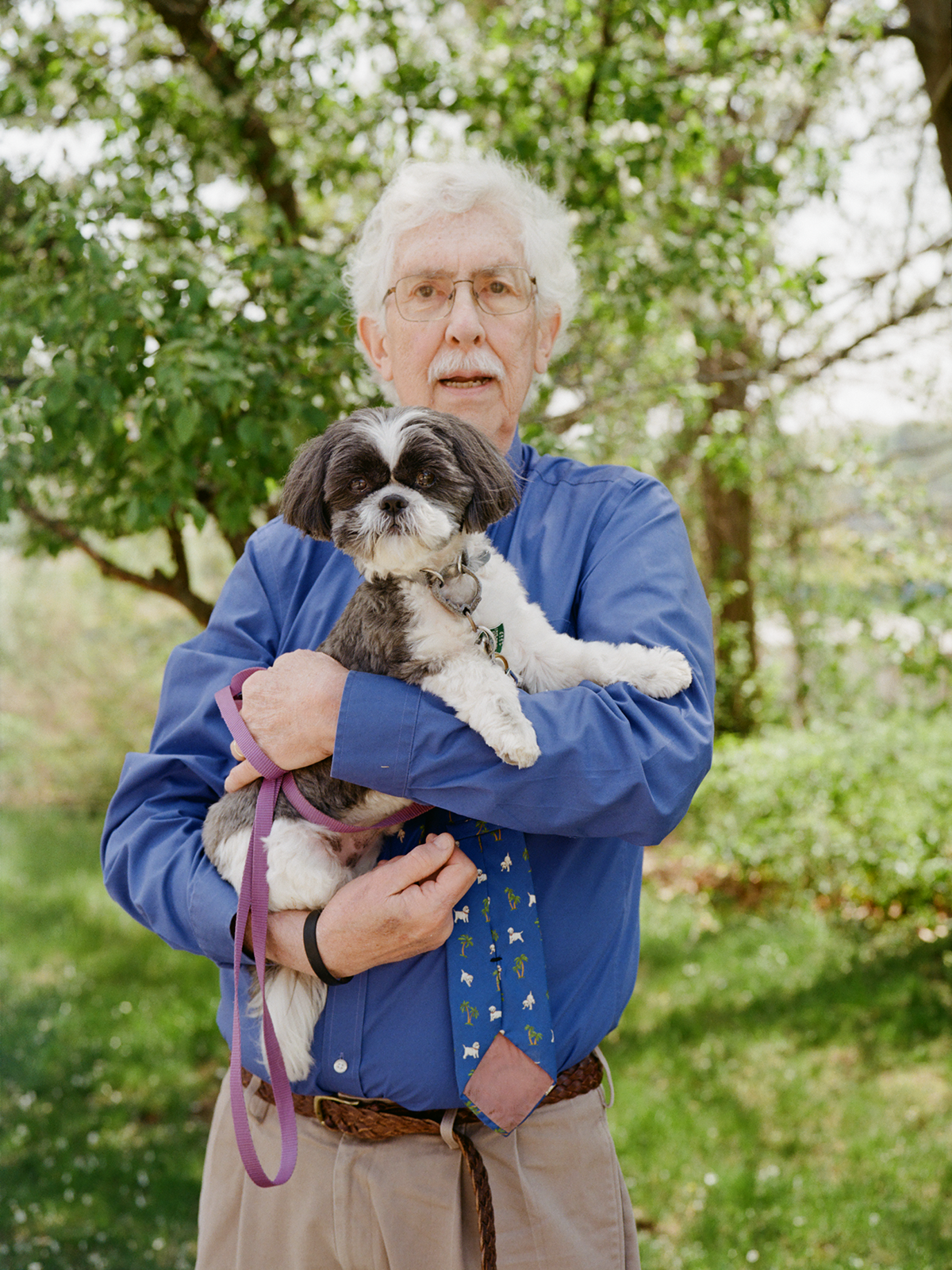 Fred Volkma in a blue shirt, outside in front of green grass and trees, holding his pet dog, who also looks at the camera.