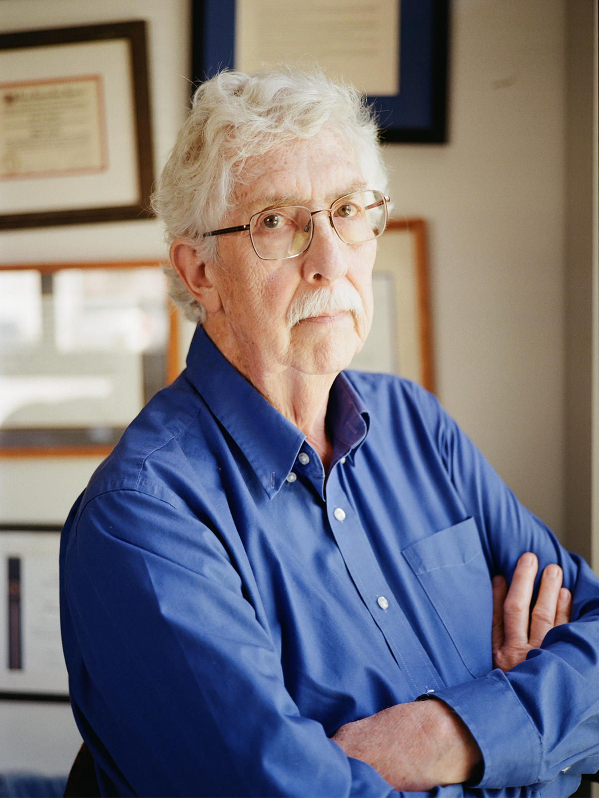 Fred Volkmar, in a blue shirt, at 3/4 length, in front of wall of framed certificates in his office, wearing a blue shirt.