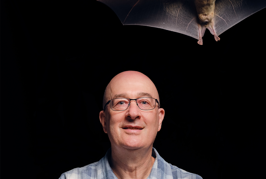 Nachum Ulanovsky sits against a black background with one bat in his hands and another with its wings spread above his head.
