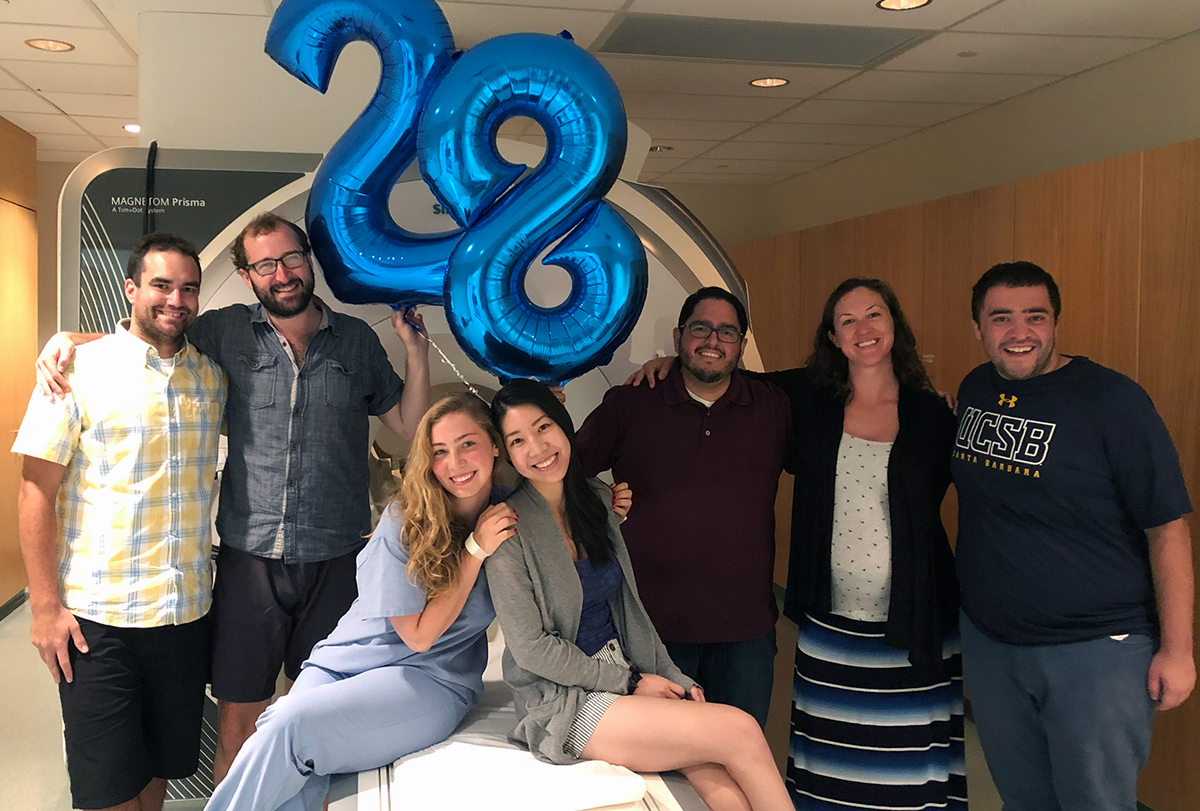 Laura Pritschet in blue scrubs is at the center of a photograph of the study team that helped with data collection (MRI, blood processing, cognitive assessments), posing on a scanner, with helium balloons in the shape of a 28.