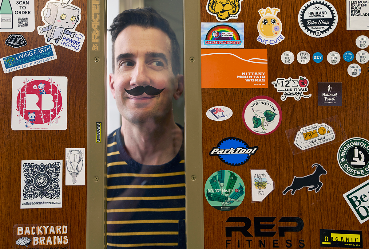 Kieth Hengen looks through a small window, aligning his face with a fancy moustache sticker and rolling his eyes comically to the side.