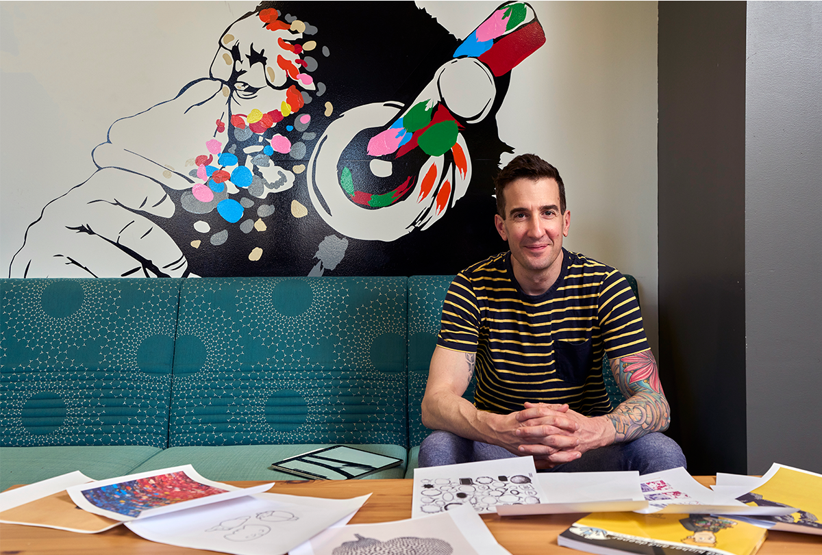 Keith Hengen in front of a wall mural in his lab, with drawings scattered on the table in front of him.