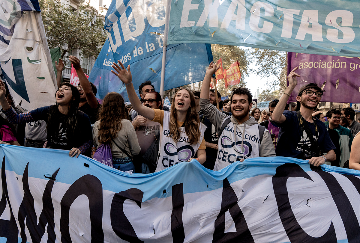 Manifestantes que gritan y corean llevan una gran pancarta de tela frente a ellos, y se aprecian otras pancartas de tela más pequeñas sobre sus cabezas.