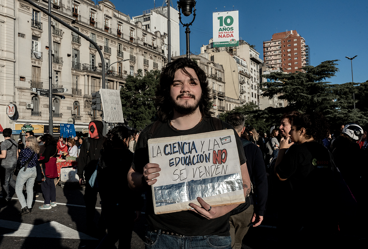 Un joven de cabello oscuro y bigote sostiene un cartel hecho a mano en las calles de Buenos Aires.
