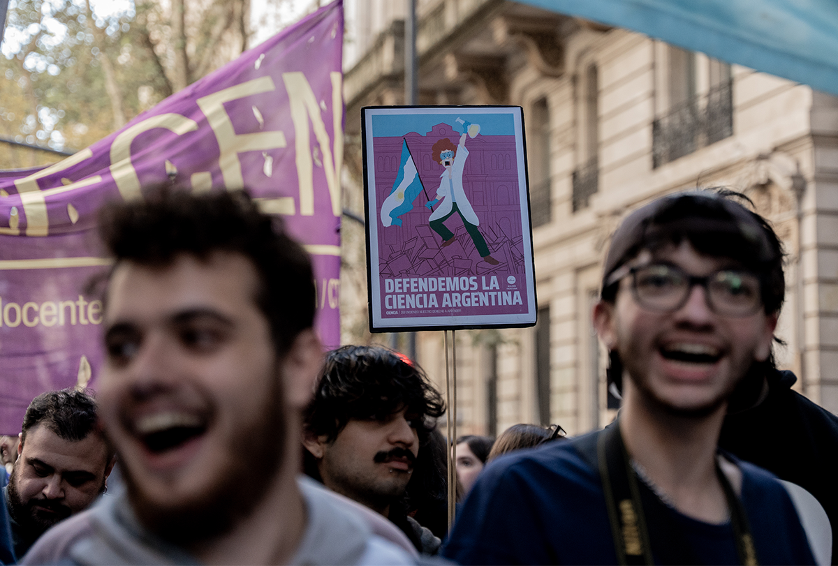 Manifestantes portan pancartas y corean consignas en las calles de Buenos Aires: dos jóvenes aparecen en primer plano con la boca abierta y, detrás de ellos, el foco de atención recae sobre una pancarta de protesta acerca de la importancia de la financiación de la ciencia.