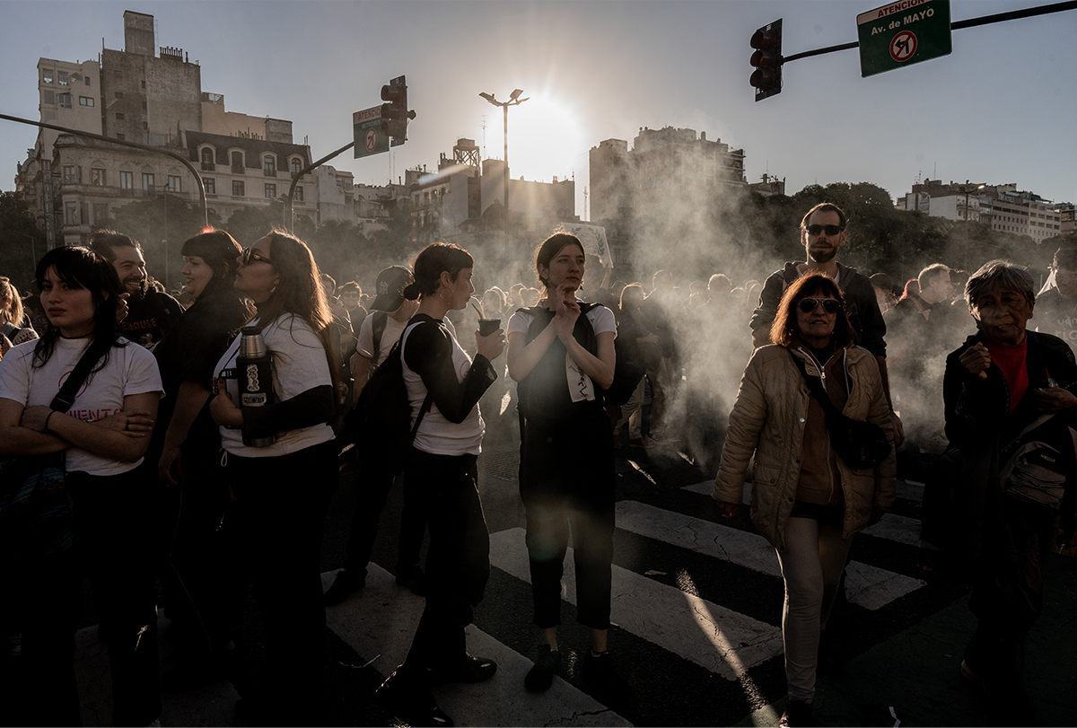 Se observa a un grupo de mujeres manifestantes a contraluz, parcialmente silueteadas, proyectando oscuras sombras. El humo de un vendedor de comida cercano contribuye a la atmósfera.