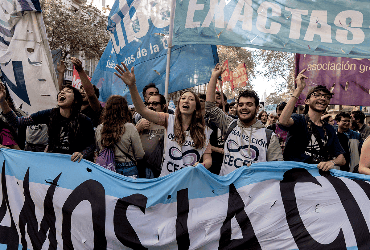 Manifestantes que gritam e entoam palavras de ordem carregam uma grande faixa de tecido à sua frente, e outras faixas de tecido menores são visíveis acima de suas cabeças.