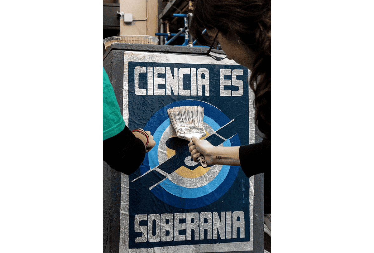 Mãos estão colando um cartaz em um espaço público com um pincel; o cartaz traz a frase "Ciencia es soberania" — que significa "Ciência é soberania" —, um slogan popular na Argentina.