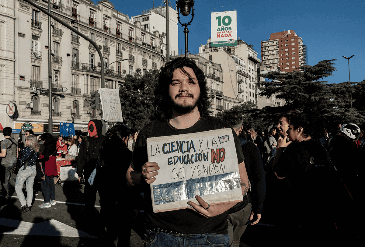 Um jovem de cabelos escuros e bigode segura uma placa feita à mão nas ruas de Buenos Aires.