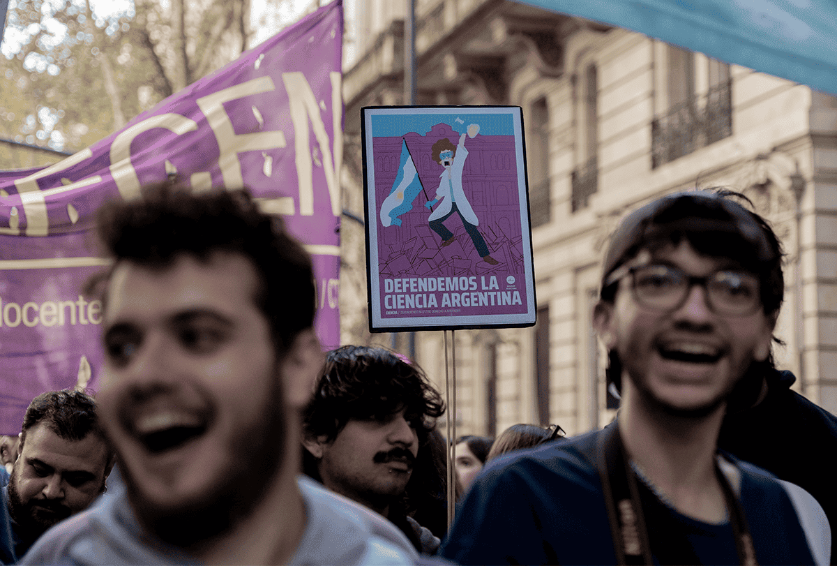 Manifestantes carregam cartazes e entoam palavras de ordem nas ruas de Buenos Aires: dois jovens aparecem em primeiro plano com as bocas abertas e, atrás deles, o foco recai sobre um cartaz de protesto que destaca a importância do financiamento da ciência.