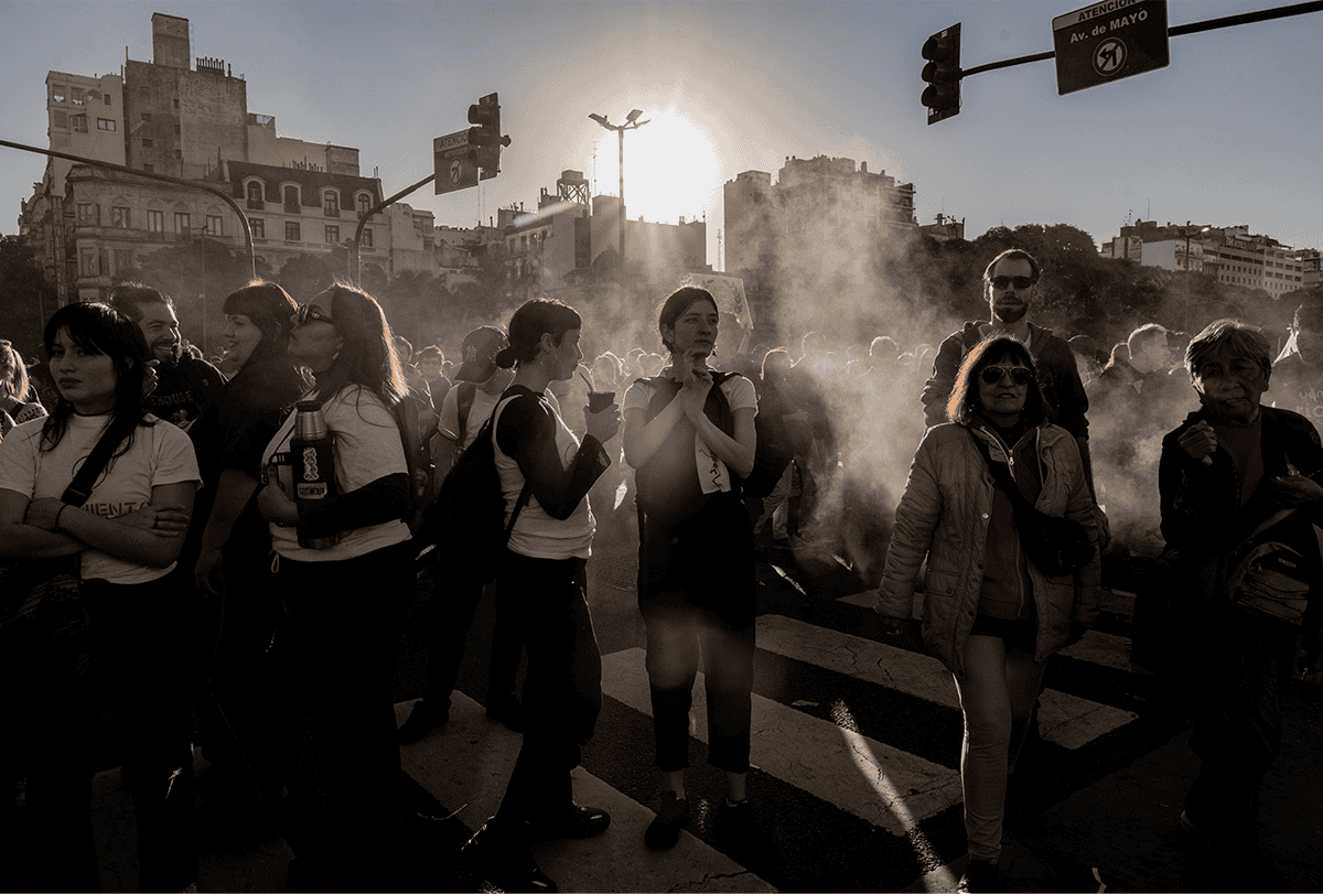 Um grupo de mulheres manifestantes é visto contra o sol, em silhueta parcial, projetando sombras escuras. A fumaça de um vendedor de comida próximo contribui para a atmosfera.