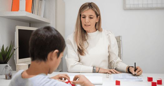 A boy manipulates blocks at a table while sitting across from a woman taking notes